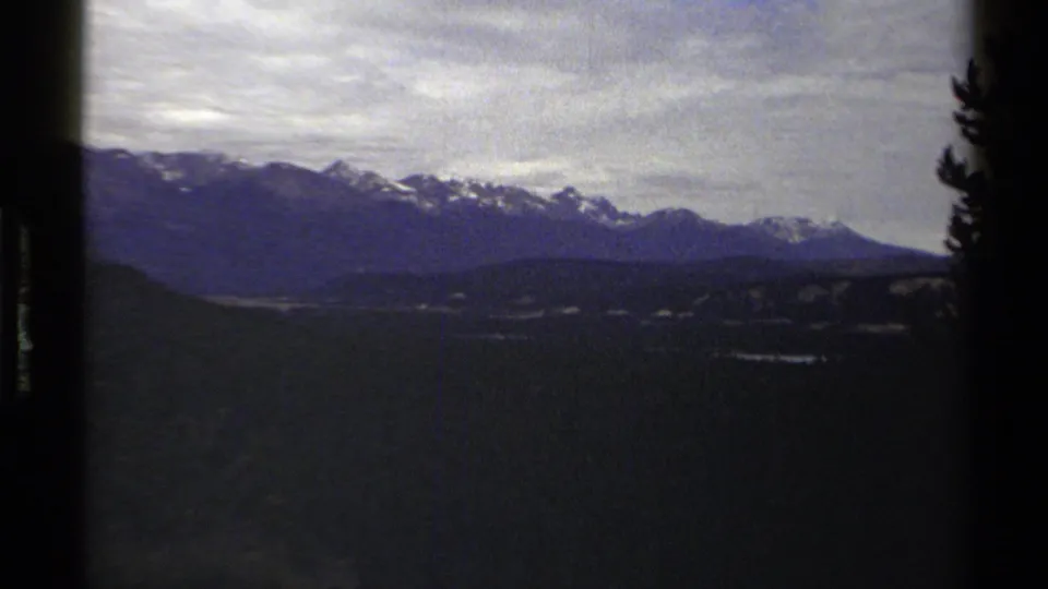 1979 8mm travel frame with British Columbia mountain peaks, cloudy sky, dark foreground terrain, and a tree silhouette at the right edge.