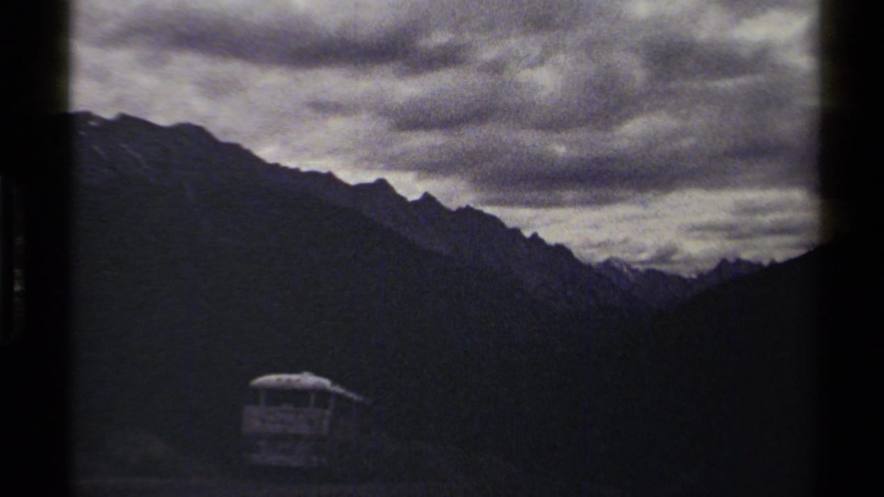 British Columbia 8mm travel frame showing a small bus on a mountain road with steep dark ridges and heavy cloud cover.
