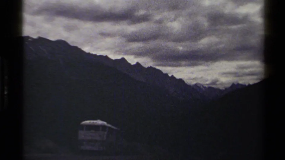 British Columbia 8mm travel frame showing a small bus on a mountain road with steep dark ridges and heavy cloud cover.