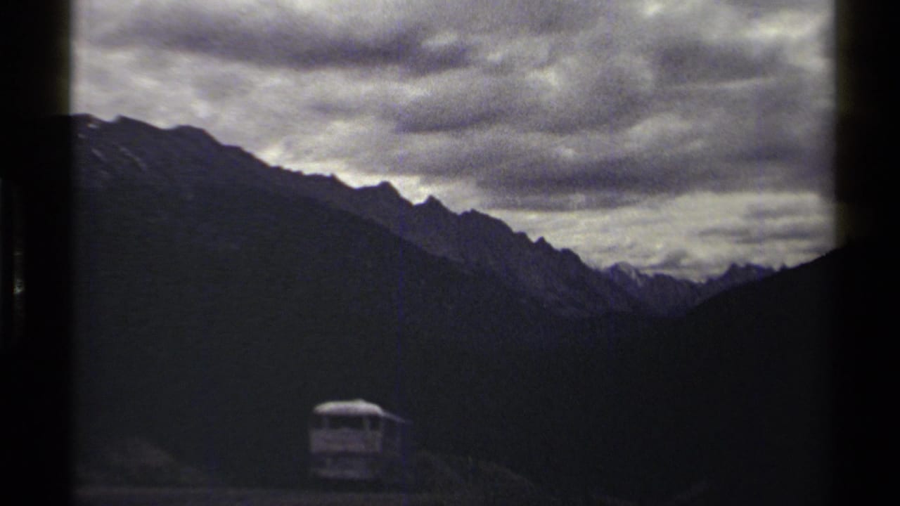 1979 8mm British Columbia mountain view with slate clouds, jagged ridges, dark valley slopes, and a small distant vehicle.
