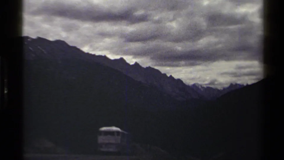 1979 8mm British Columbia mountain view with slate clouds, jagged ridges, dark valley slopes, and a small distant vehicle.