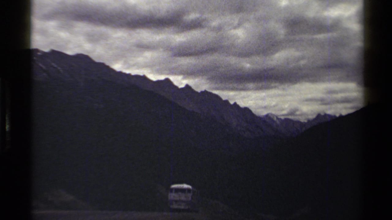 British Columbia mountain scene on 8mm film, showing a small distant vehicle below dark ridges and layered storm clouds.