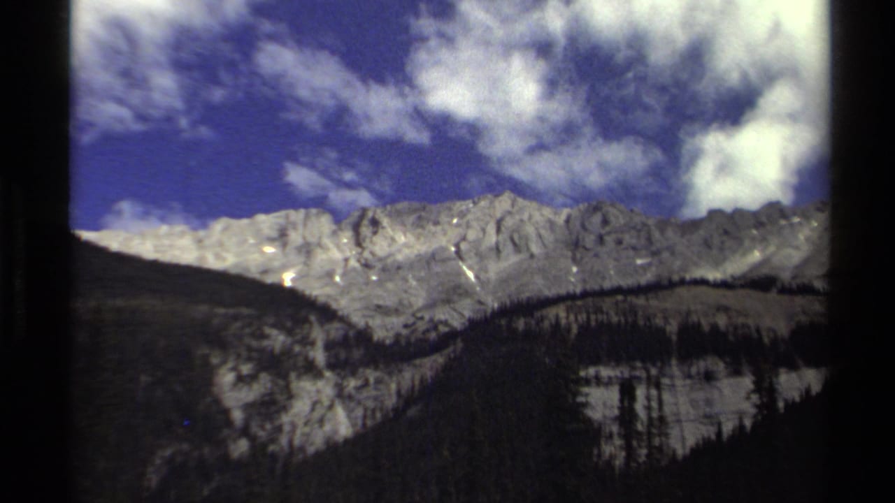 1979 8mm British Columbia landscape with cobalt sky, bright clouds, pale rocky peaks, and dark evergreen forest below.
