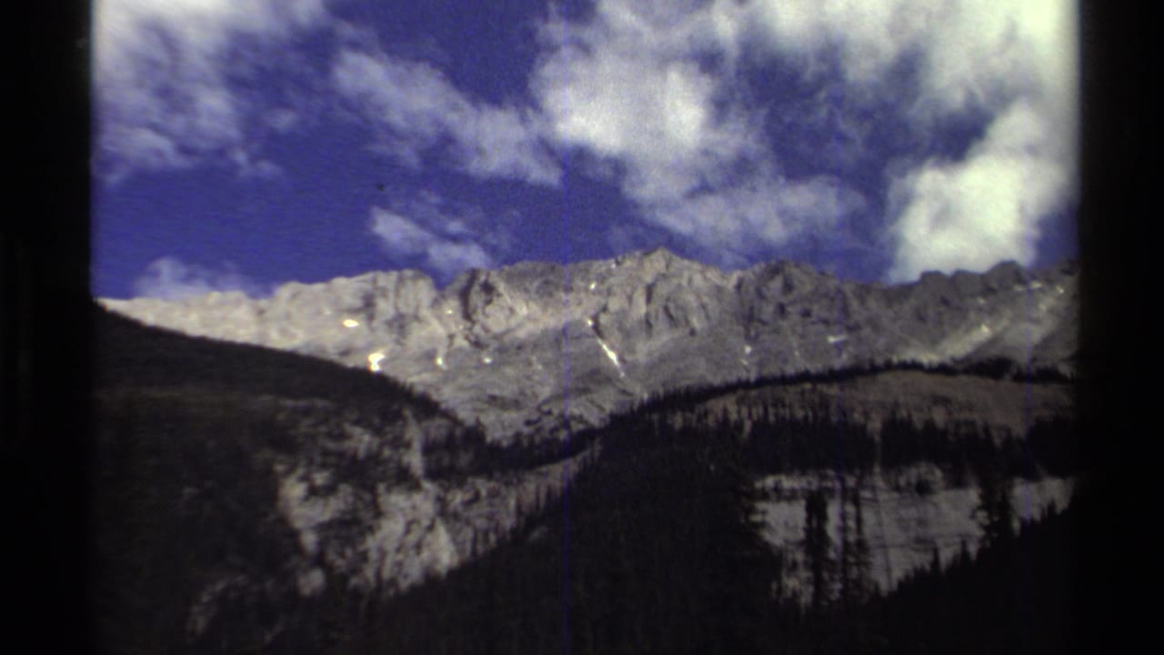 British Columbia mountain view on 8mm film, with bright clouds, pale ridgelines, and dark evergreen-covered slopes.