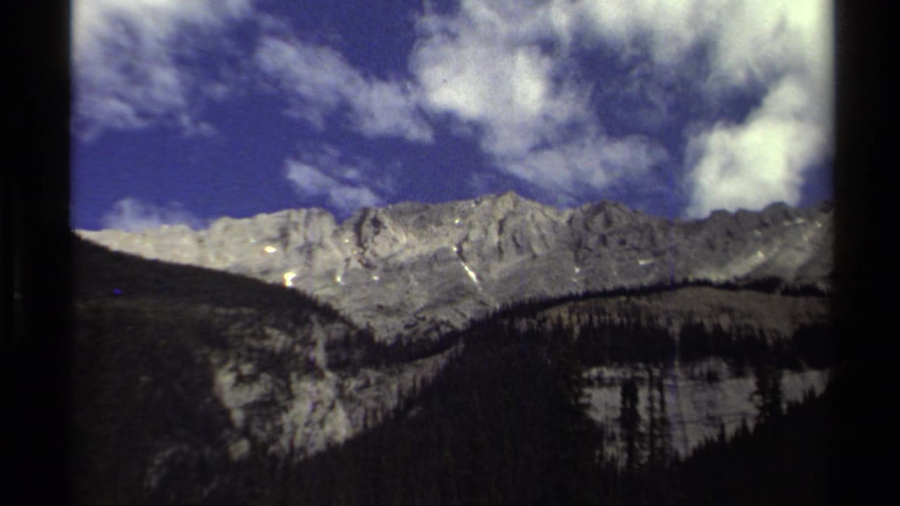 1979 8mm home-movie frame of a rugged British Columbia mountain range beneath a deep blue sky and white clouds.