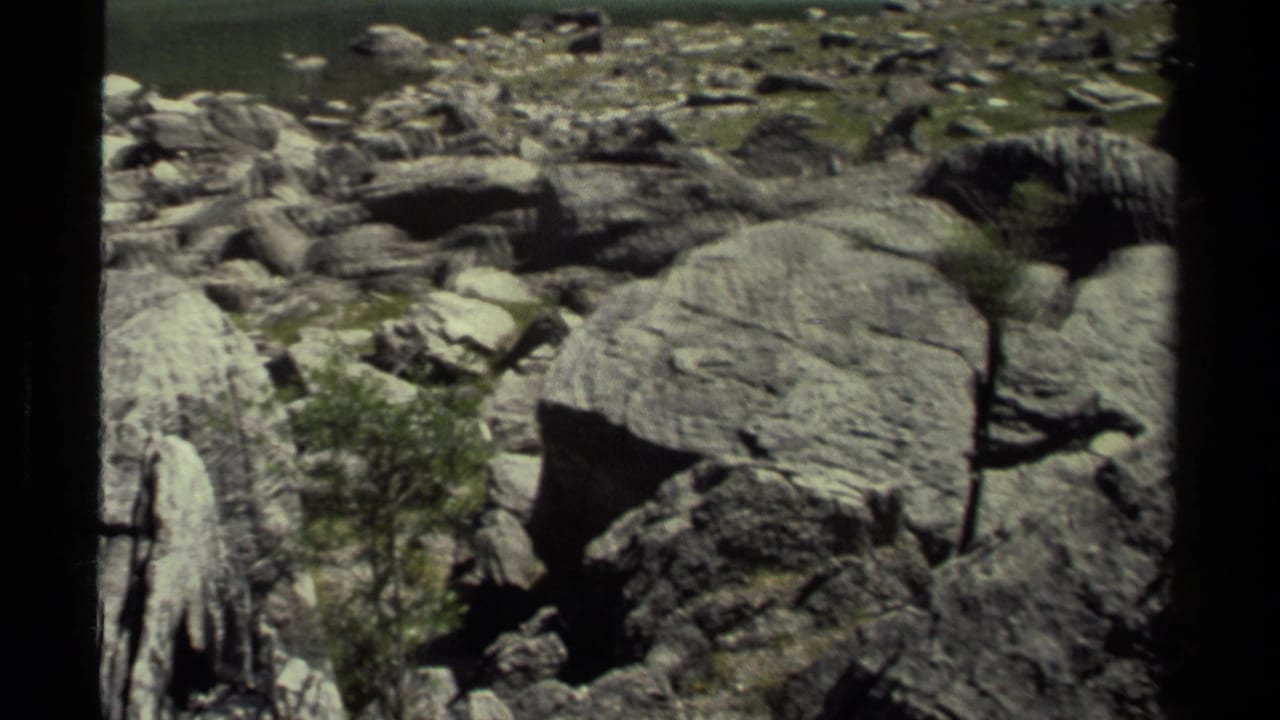 1979 8mm British Columbia frame of rocky ground with a broad flat boulder, moss patches, and low shrubs or small trees.