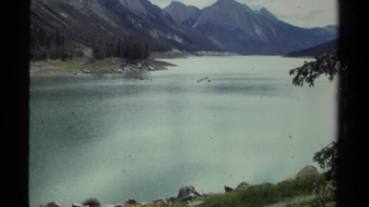 1979 8mm British Columbia lake view with turquoise water, forested mountain slopes, shoreline rocks, and distant peaks under a pale sky.