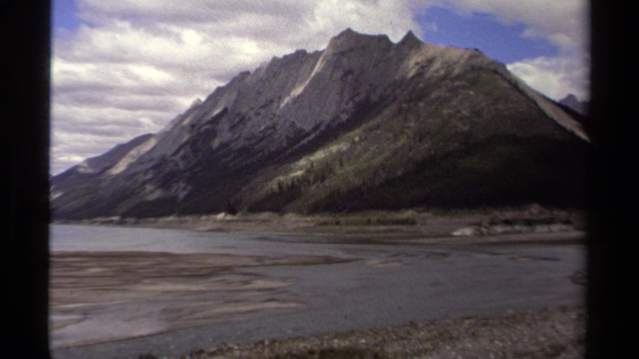 British Columbia mountain landscape from a 1979 8mm home movie, with water and gravel shore below a partly cloudy sky.