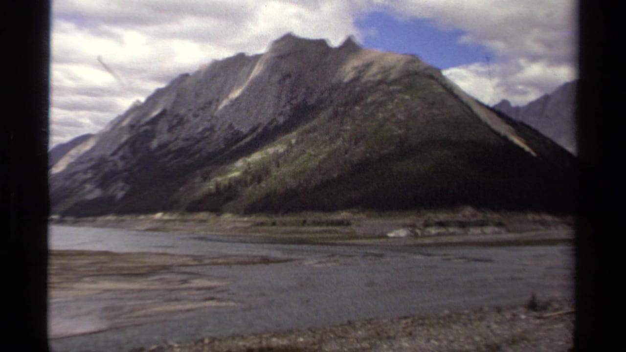 1979 British Columbia 8mm frame showing rugged mountains, a forested slope, and a wide river or lake foreground.