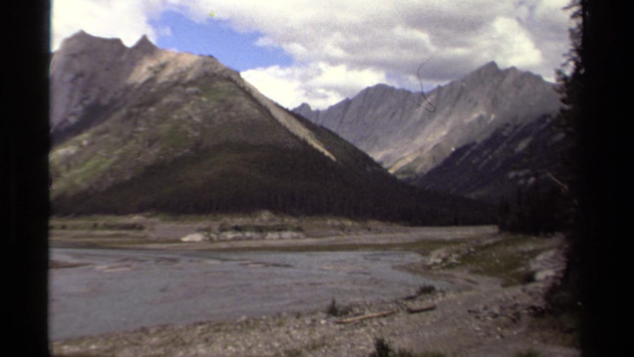1979 British Columbia 8mm frame of a mountain range across a river valley, with dark trees entering the right edge.