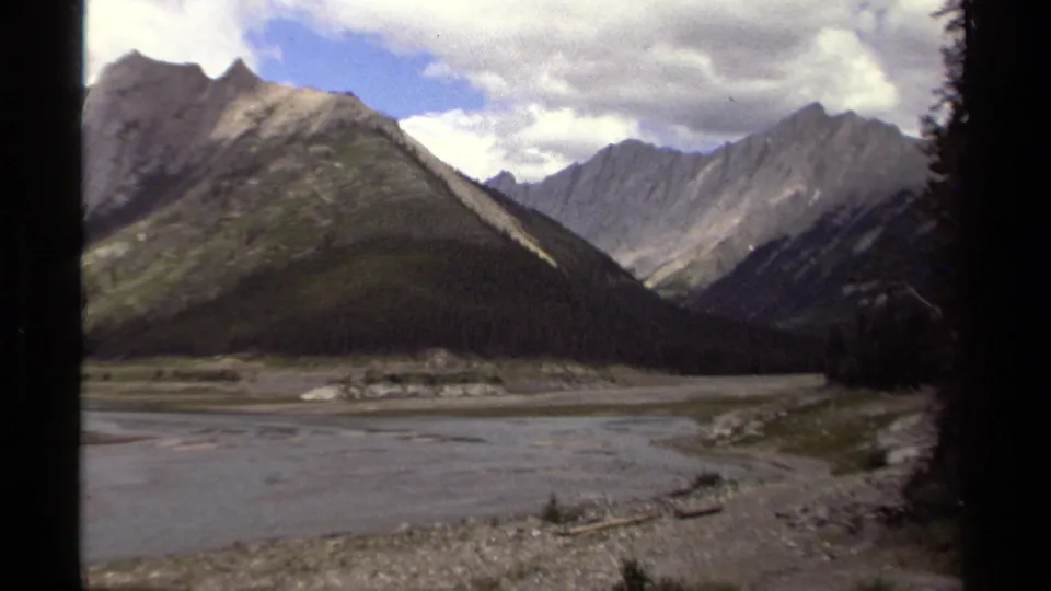 1979 8mm British Columbia view of a broad riverbed running through a rugged mountain valley under broken clouds.
