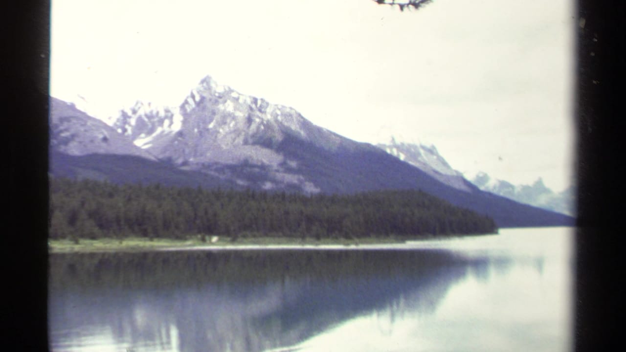 1979 8mm British Columbia lake scene with snow-capped mountain peaks, evergreen shoreline, and still water reflections.