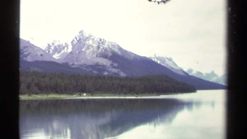 1979 8mm British Columbia lake scene with snow-capped mountain peaks, evergreen shoreline, and still water reflections.
