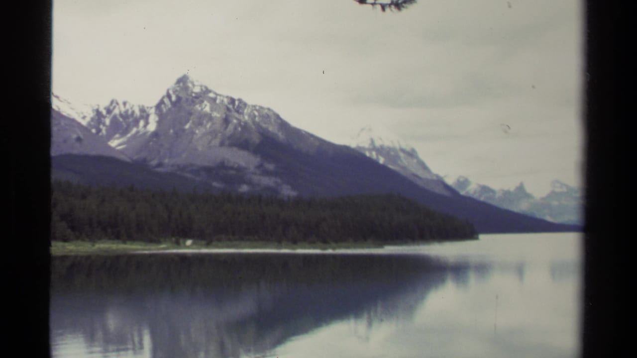 Vintage 8mm British Columbia landscape showing layered mountains, dark evergreen forest, and muted reflections on a still lake.