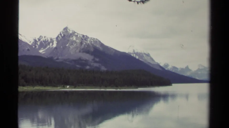 Vintage 8mm British Columbia landscape showing layered mountains, dark evergreen forest, and muted reflections on a still lake.