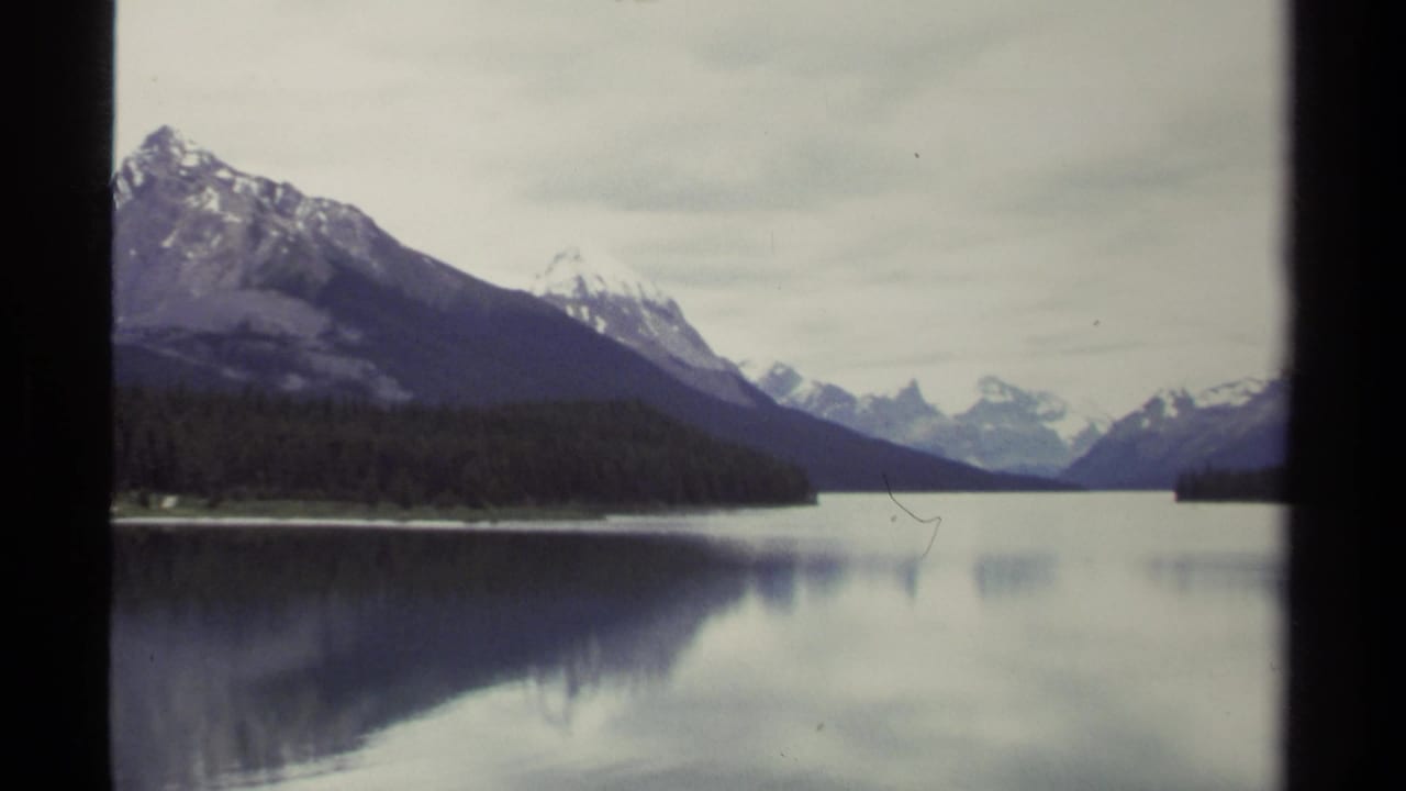 1979 8mm view of a British Columbia lake with forested shoreline, snow-capped mountains, and soft reflections under overcast sky.
