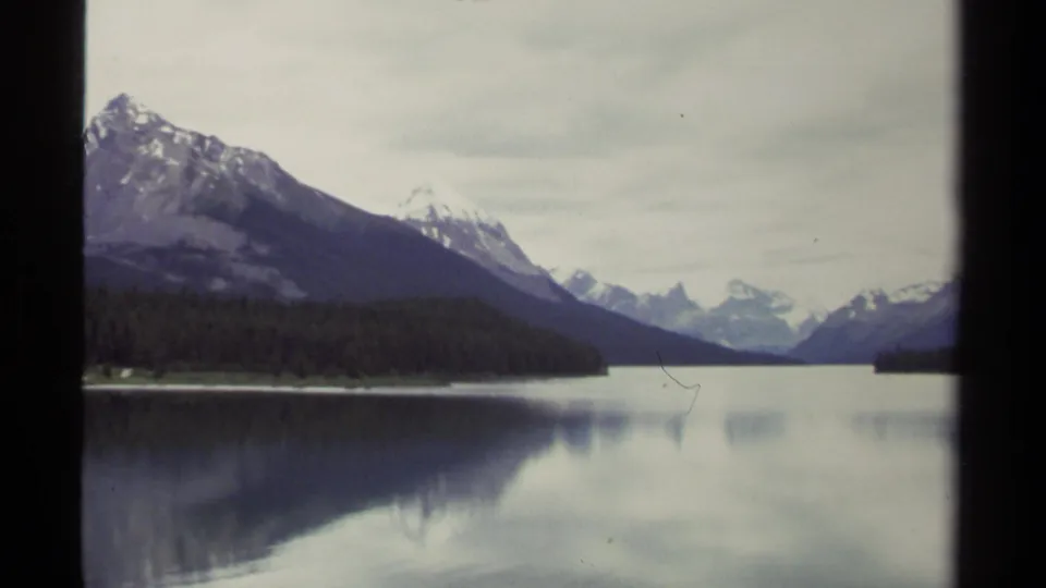 1979 8mm view of a British Columbia lake with forested shoreline, snow-capped mountains, and soft reflections under overcast sky.
