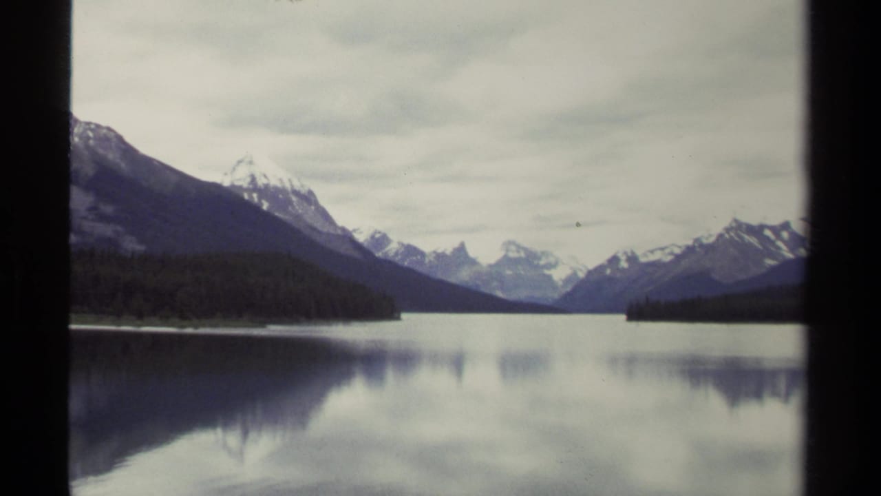 British Columbia mountain lake scene on 8mm film, with dark forest edges, distant snowy peaks, and still water reflecting the sky.