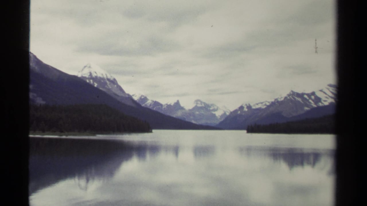 1979 home-movie frame of a British Columbia lake, showing snowy mountains beyond evergreen forest and muted reflections on calm water.