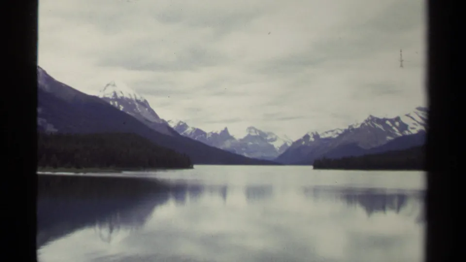 1979 home-movie frame of a British Columbia lake, showing snowy mountains beyond evergreen forest and muted reflections on calm water.