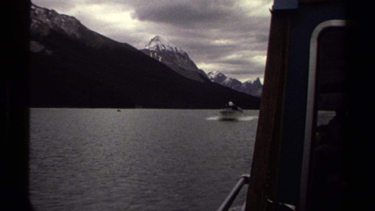 1979 British Columbia 8mm view from a boat of a small motorboat crossing a mountain lake below snow-capped peaks.