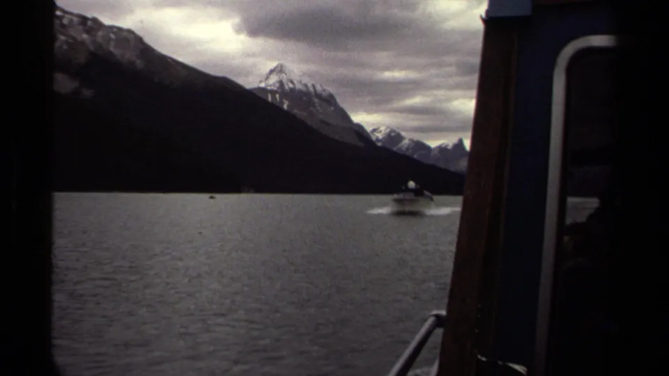 1979 British Columbia 8mm view from a boat of a small motorboat crossing a mountain lake below snow-capped peaks.