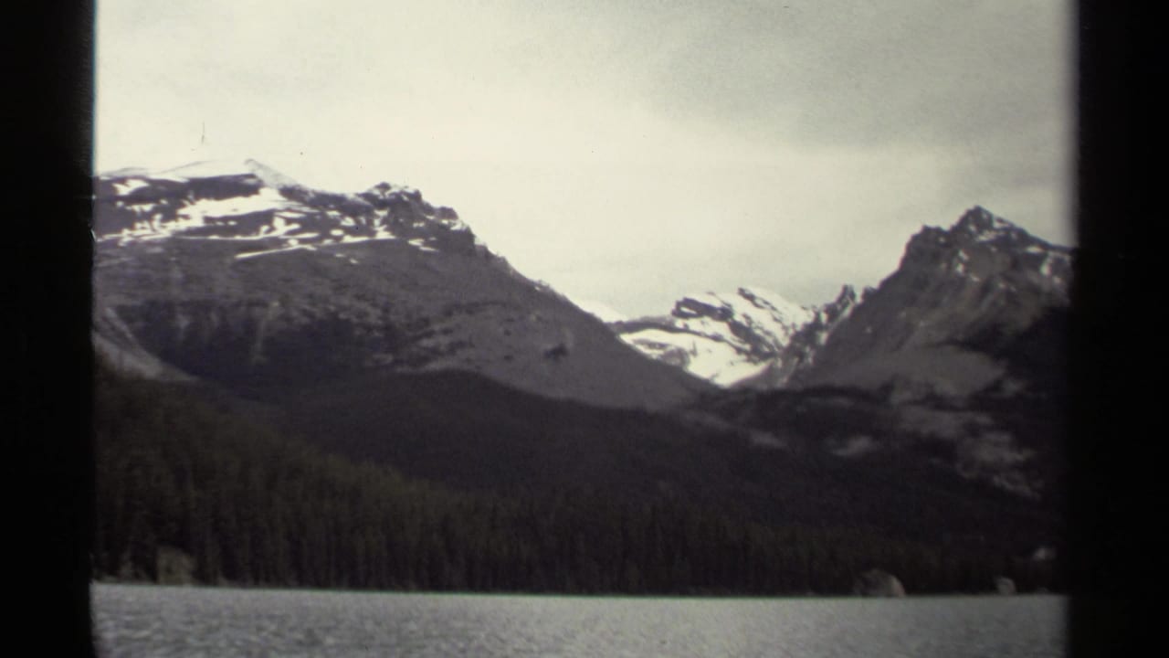 British Columbia mountain landscape on 8mm film, showing lake water, evergreen shoreline, and snow-streaked peaks under pale cloud.