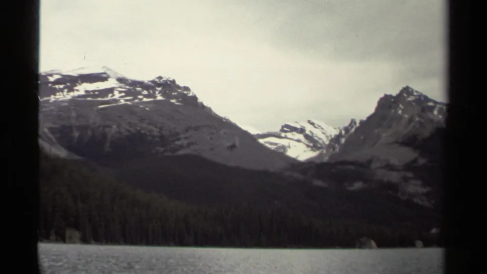 British Columbia mountain landscape on 8mm film, showing lake water, evergreen shoreline, and snow-streaked peaks under pale cloud.