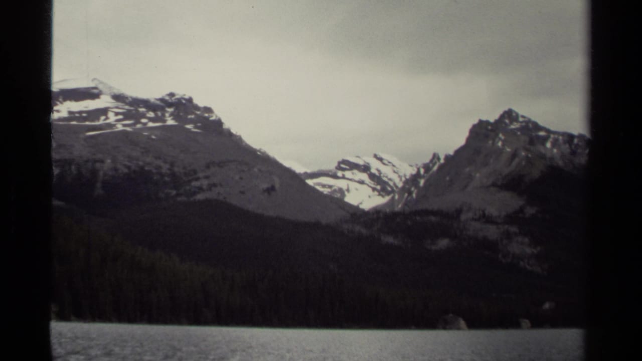 1979 8mm view of British Columbia mountains rising above evergreen forest and a calm lake under an overcast sky.