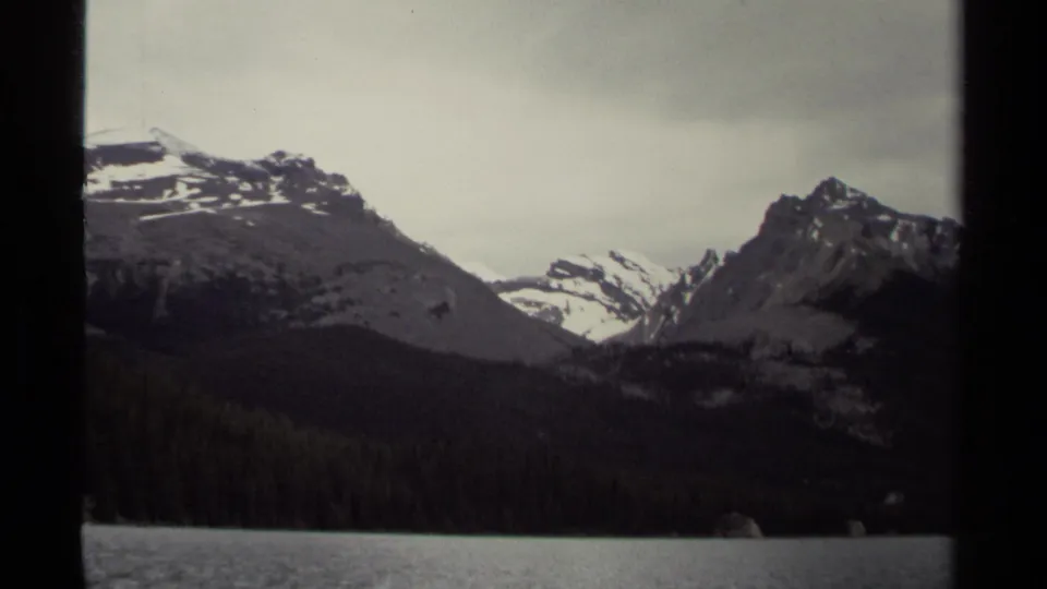 1979 8mm view of British Columbia mountains rising above evergreen forest and a calm lake under an overcast sky.