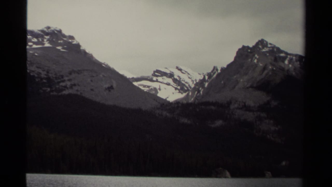 British Columbia mountain landscape on 8mm film, with snowy ridges, dark forest, and lake shoreline visible through frame borders.