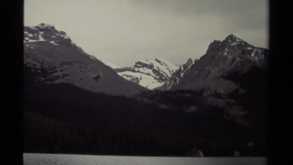 British Columbia mountain landscape on 8mm film, with snowy ridges, dark forest, and lake shoreline visible through frame borders.