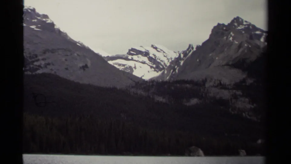 1979 home-movie frame of steep British Columbia mountain terrain, with snow patches, dark forest, and a calm lake edge.