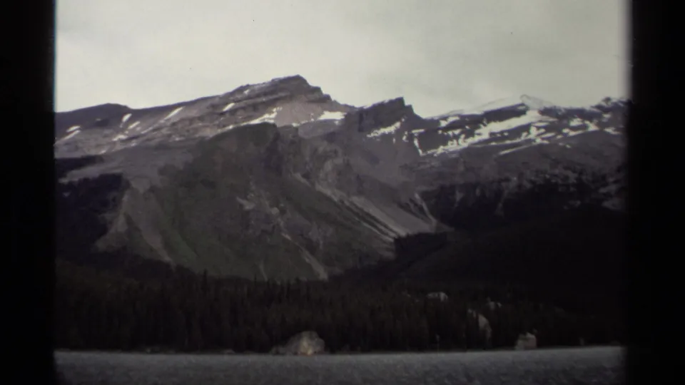 British Columbia 8mm landscape frame showing a rough rocky hillside, scattered snow, forest, and lake water.