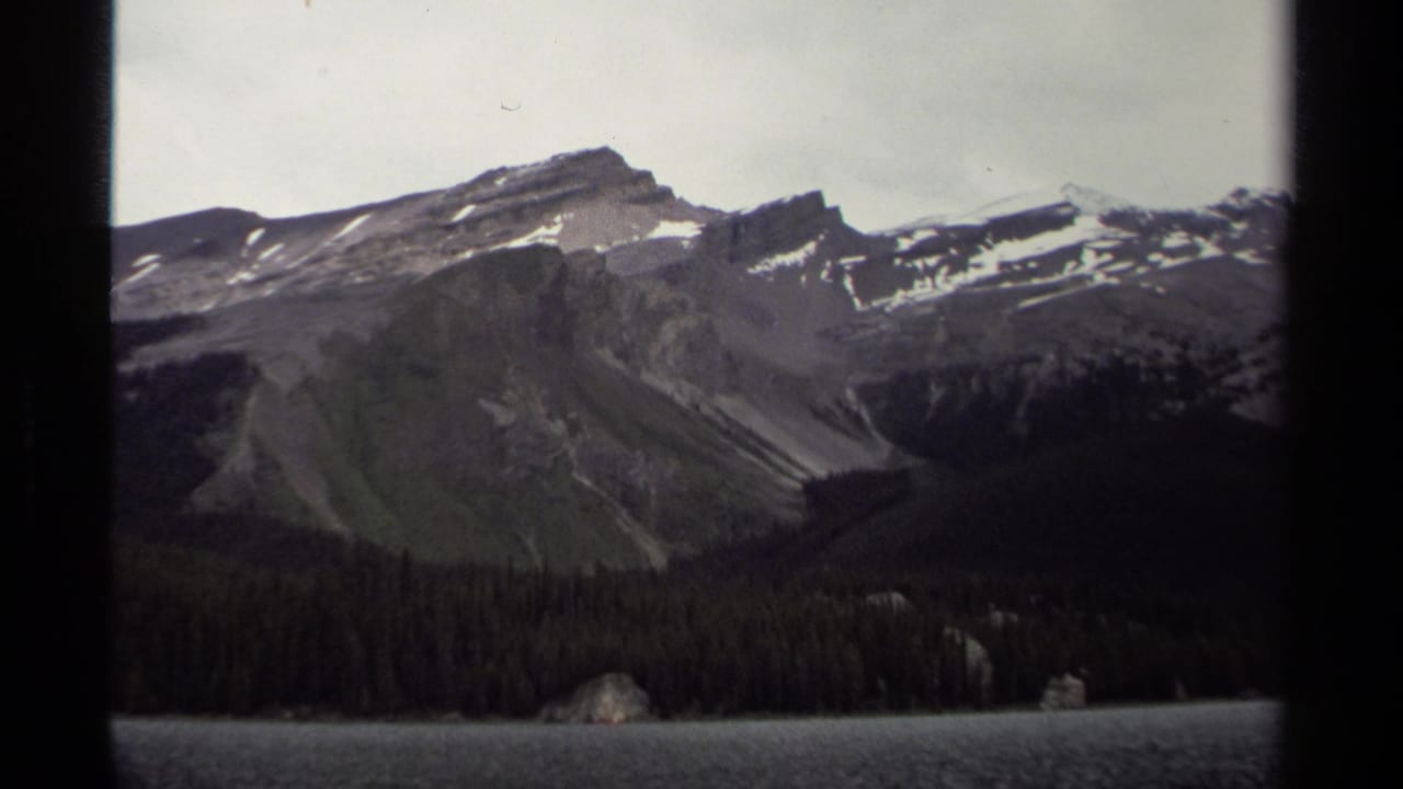 1979 8mm view of a British Columbia mountain ridge, evergreen shoreline, and calm lake under pale overcast light.