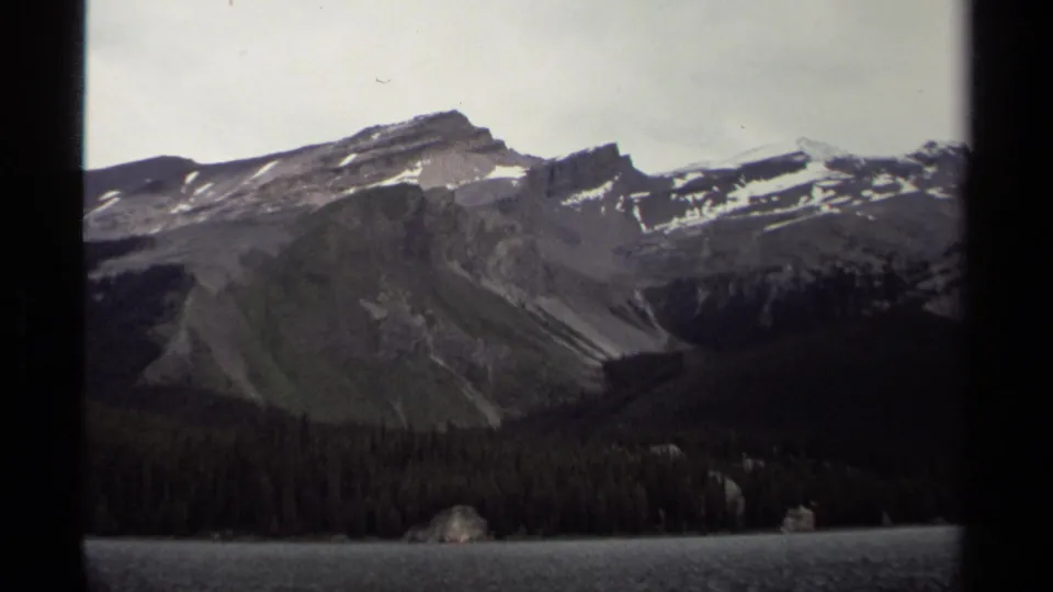 1979 8mm view of a British Columbia mountain ridge, evergreen shoreline, and calm lake under pale overcast light.