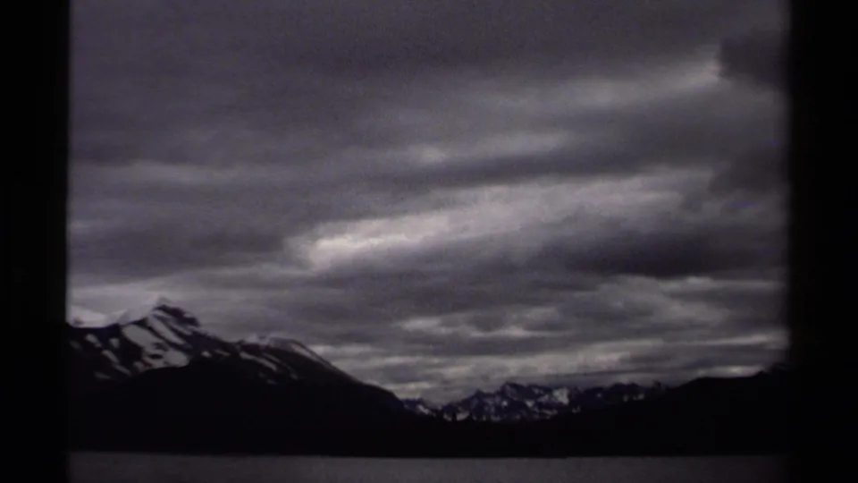 A dark 8mm frame shows a British Columbia lake and distant snow-patched mountains below layered overcast clouds.