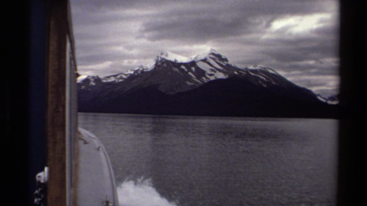 Boat wake on a lake with snow-capped mountains under a cloudy sky.