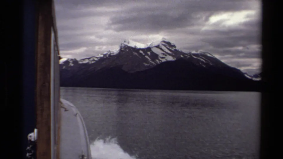 1979 8mm British Columbia boat-view footage showing lake water, white wake, dark shoreline, and snow-capped mountains beneath heavy clouds.