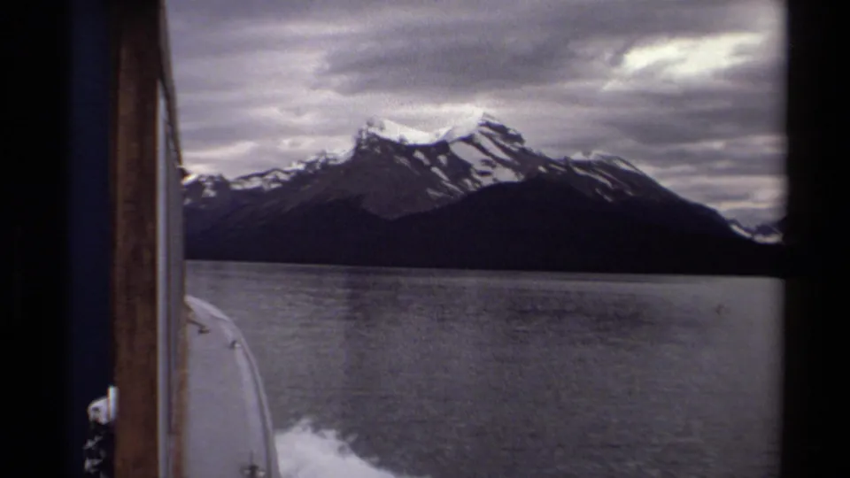 British Columbia lake scenery from a moving boat, with a dark mountain range, snow on peaks, gray clouds, and 8mm film borders.