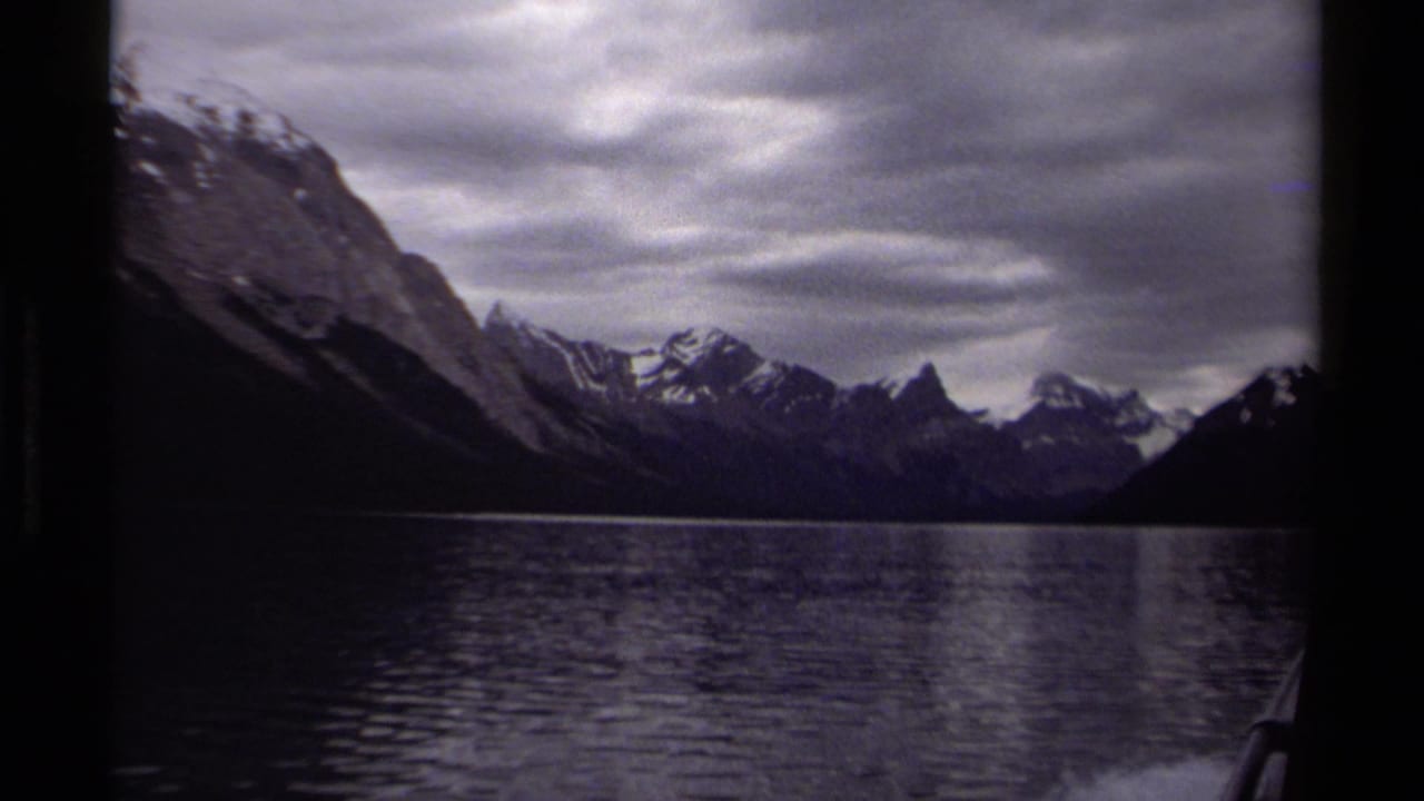 1979 British Columbia lake passage with snow-dusted peaks, calm rippling water, and a dark boat-edge framing the view.