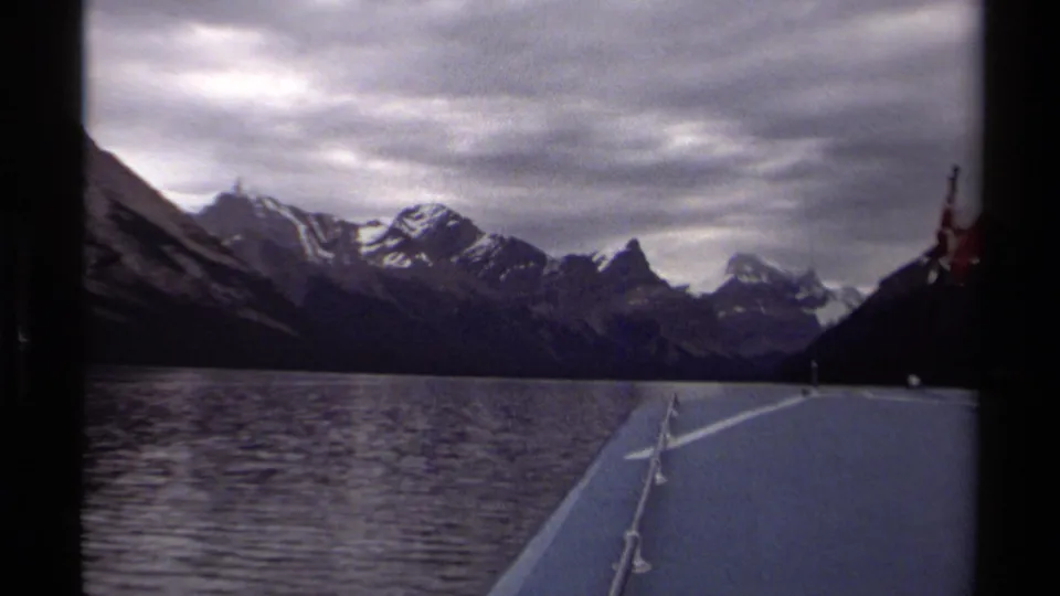 1979 8mm boat-deck view in British Columbia, showing bow railings, rippling lake water, and snow-capped mountains ahead.