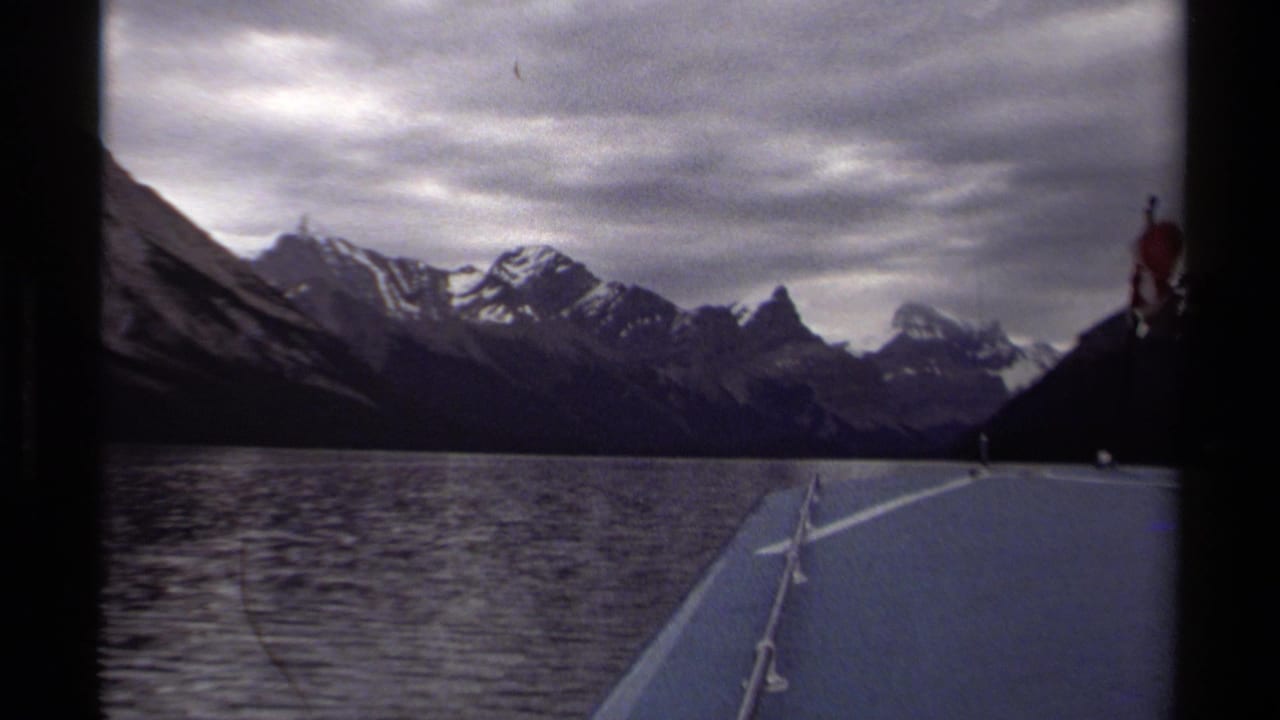 1979 British Columbia 8mm frame showing a boat bow on an alpine lake with snow-capped mountains beneath an overcast sky.