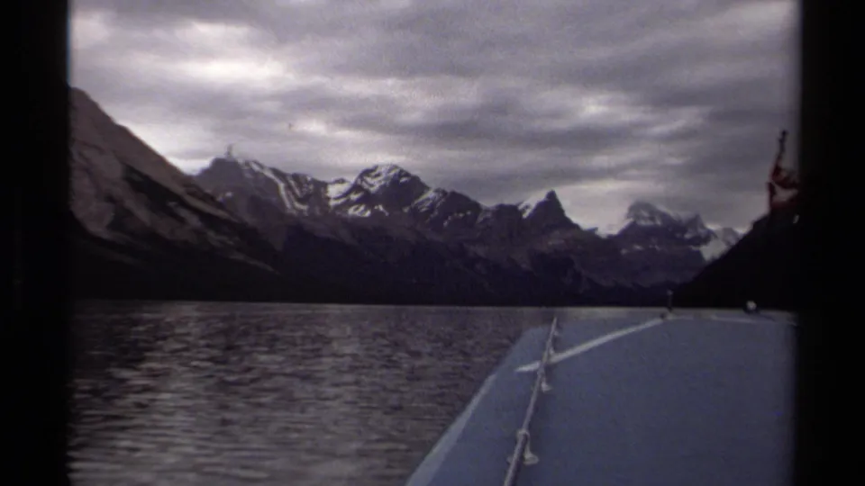 1979 British Columbia home-movie frame from a moving boat, looking across lake water to rugged snowy peaks and cloudy sky.