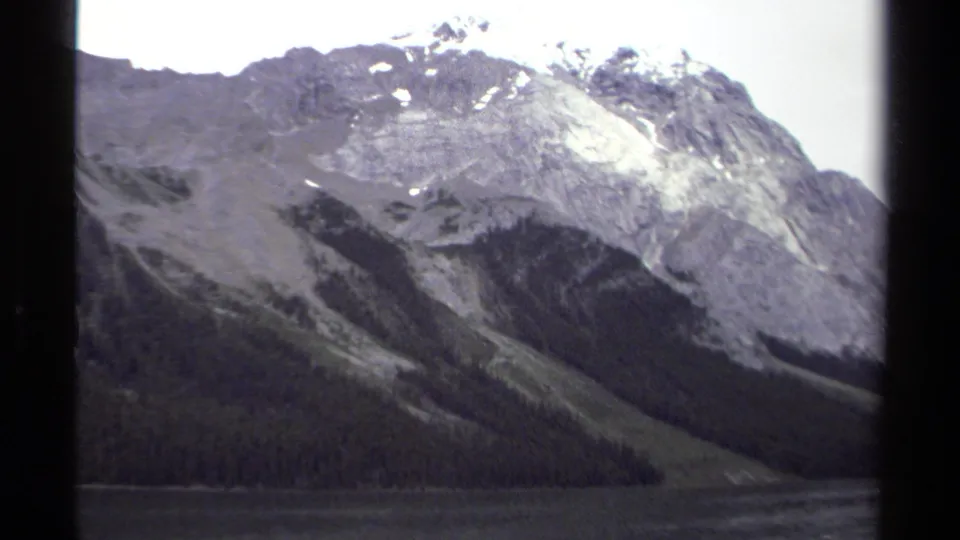 1979 British Columbia 8mm frame showing a closer alpine slope, forested shoreline, lake water, and snow high on the mountain.