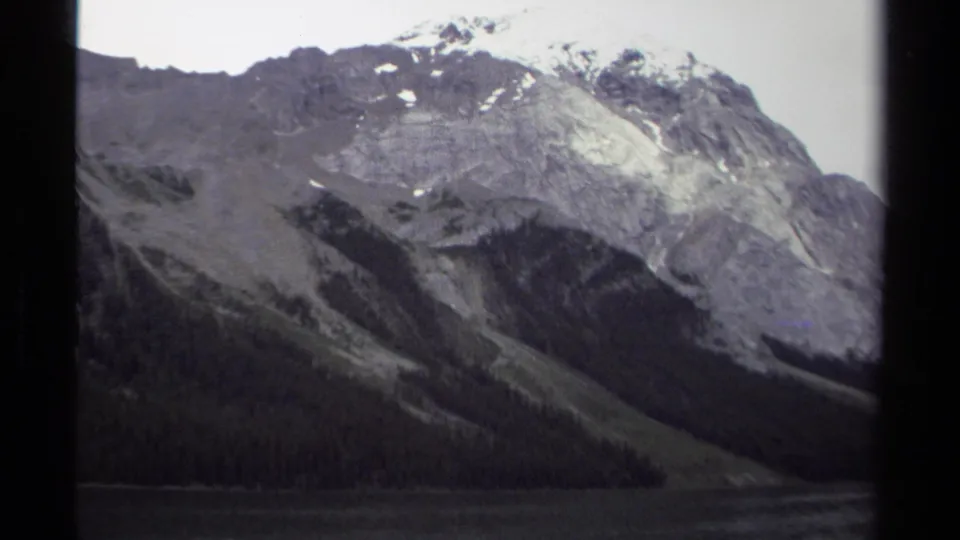 1979 British Columbia home-movie frame of a rugged mountainside above lake water, with forest at the base and snow near the ridge.