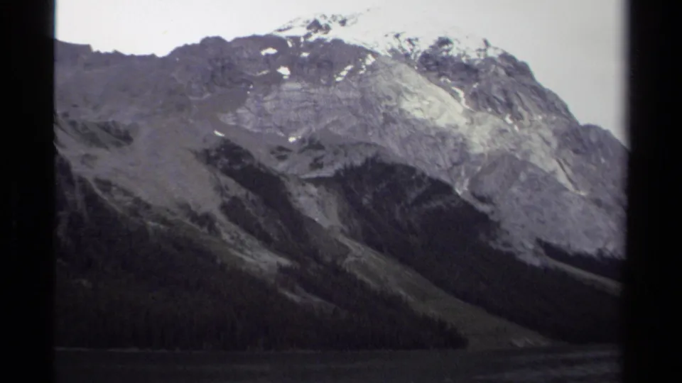 1979 British Columbia home-movie view of a snow-marked mountain rising above evergreen forest and a calm lake.