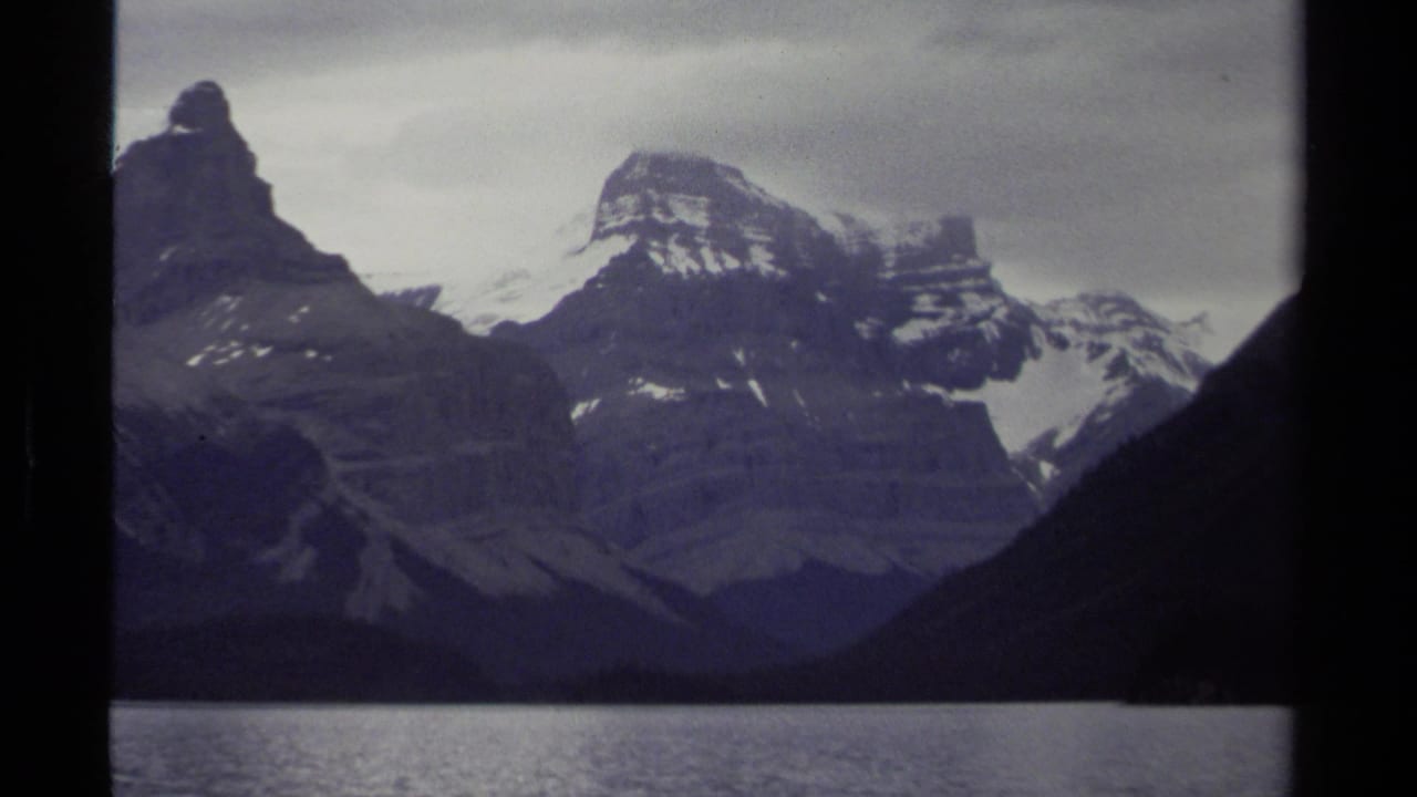 1979 British Columbia 8mm view of layered snow-capped peaks across a lake under muted overcast light.