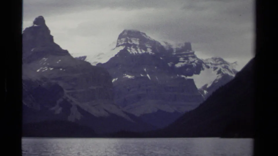 1979 British Columbia 8mm view of layered snow-capped peaks across a lake under muted overcast light.
