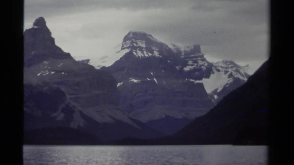 1979 British Columbia home-movie frame showing dark ridges, snow-streaked peaks, and a still lake foreground.